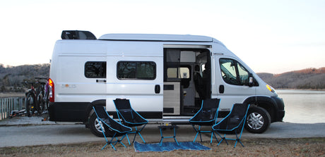 A bright Winnebago Solis RV rental parked by a lake with bikes on the rear. Camping chairs & a mat are set out.