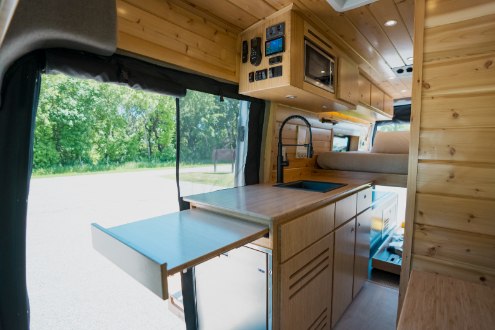 Spacious rv rental camper van interior: warm wood paneling, kitchen with sink, elevated bed, and pull-out table.