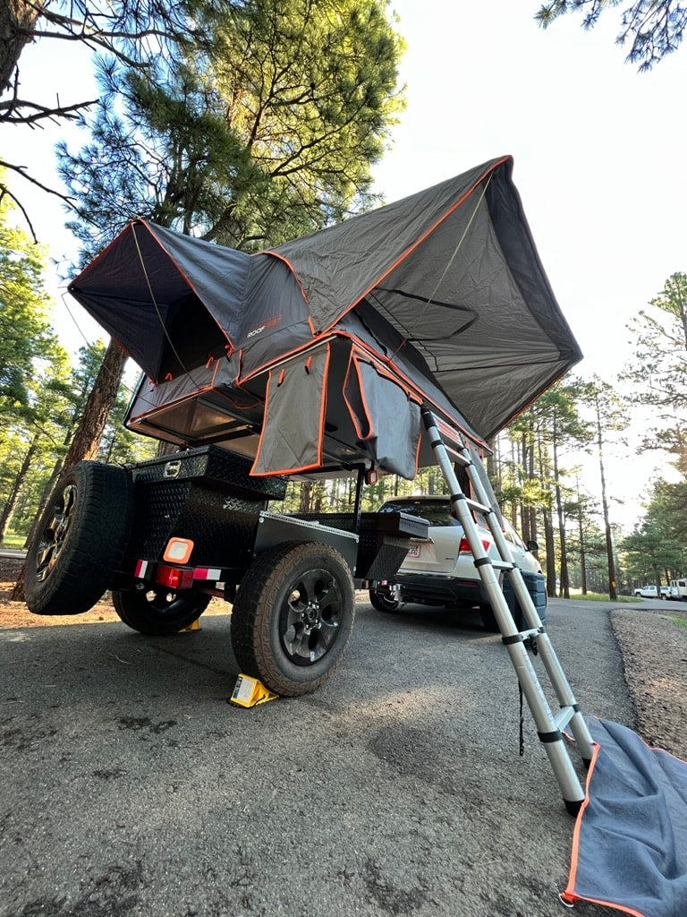 This rv rental setup features a deployed rooftop tent on an off-road camper trailer in a wooded campground. — photo 4