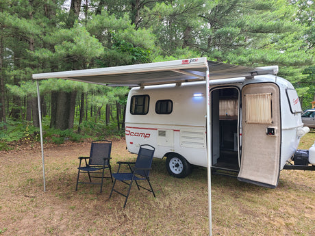 White Scamp RV rental with awning & chairs at a wooded campsite, ready for outdoor adventure.