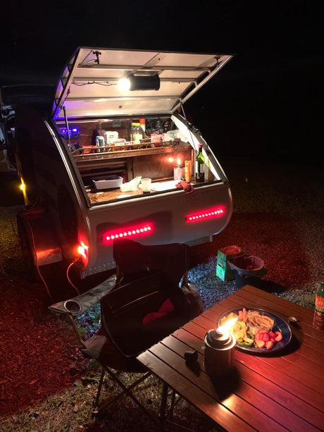 Cozy RV rental night: Teardrop camper kitchen lit up, warm glow on an outdoor meal setup. Perfect for camping!