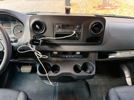 Inside an rv rental Sprinter van, this photo shows the dashboard, steering wheel, radio, cupholders, and driver's footwell.