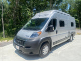 Silver camper van RV rental, ready for adventure! Parked on concrete driveway with a backdrop of green forest.