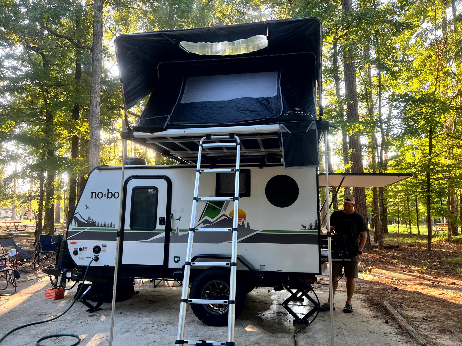 Enjoying a no-bo RV rental with a rooftop tent at a sunny campsite, complete with ladder access. A man stands nearby.