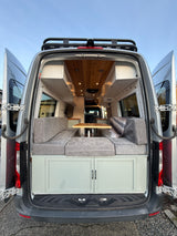 This rv rental is ready for adventure! Rear view of a custom camper van, doors open to a wood-paneled interior with gray seating.