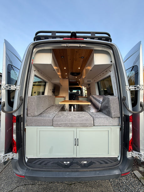 This rv rental is ready for adventure! Rear view of a custom camper van, doors open to a wood-paneled interior with gray seating.