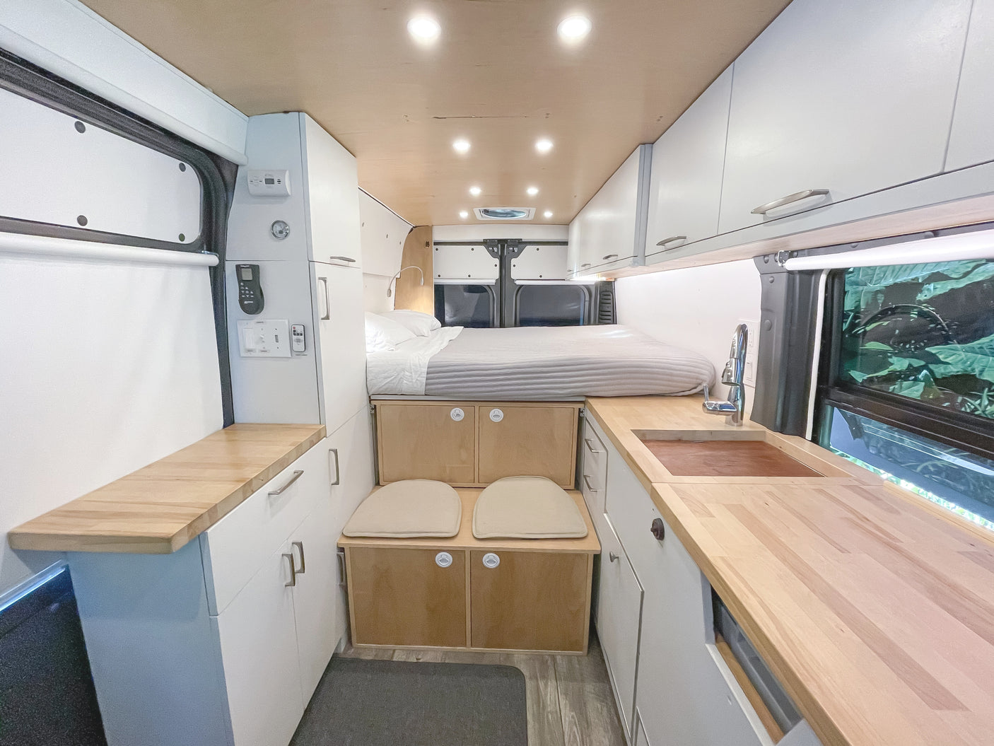 An rv rental interior with a raised bed, compact kitchen featuring a wood counter and sink, white cabinets, and small built-in seating.
