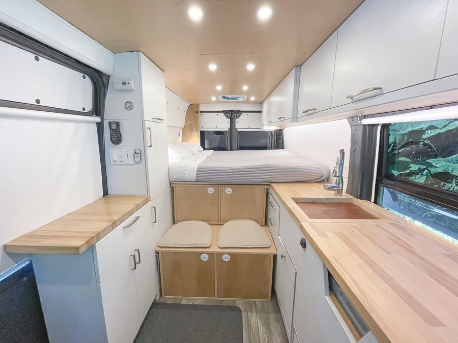 An rv rental interior with a raised bed, compact kitchen featuring a wood counter and sink, white cabinets, and small built-in seating.
