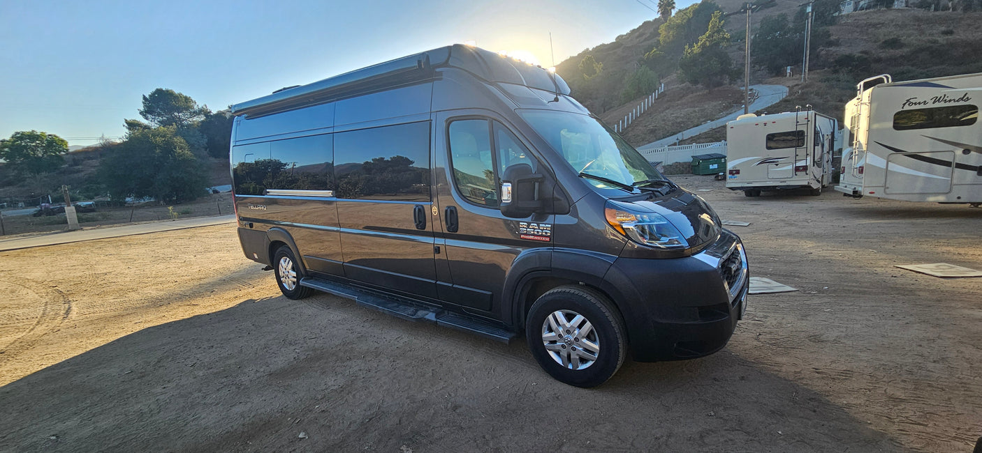 Dark grey Telluro Ram ProMaster 3500 campervan RV rental parked on dirt, with other RVs and a hillside in background.