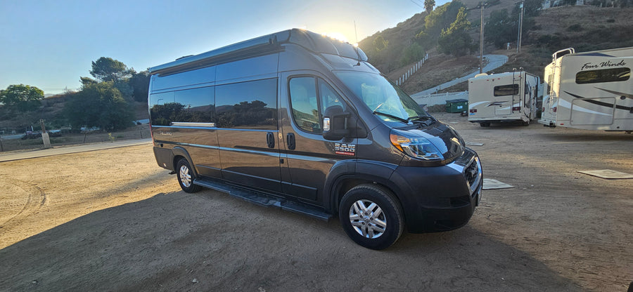Dark grey Telluro Ram ProMaster 3500 campervan RV rental parked on dirt, with other RVs and a hillside in background.
