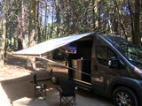 A dark grey rv rental camper van with an awning and outdoor TV is parked in a shaded forest campsite.