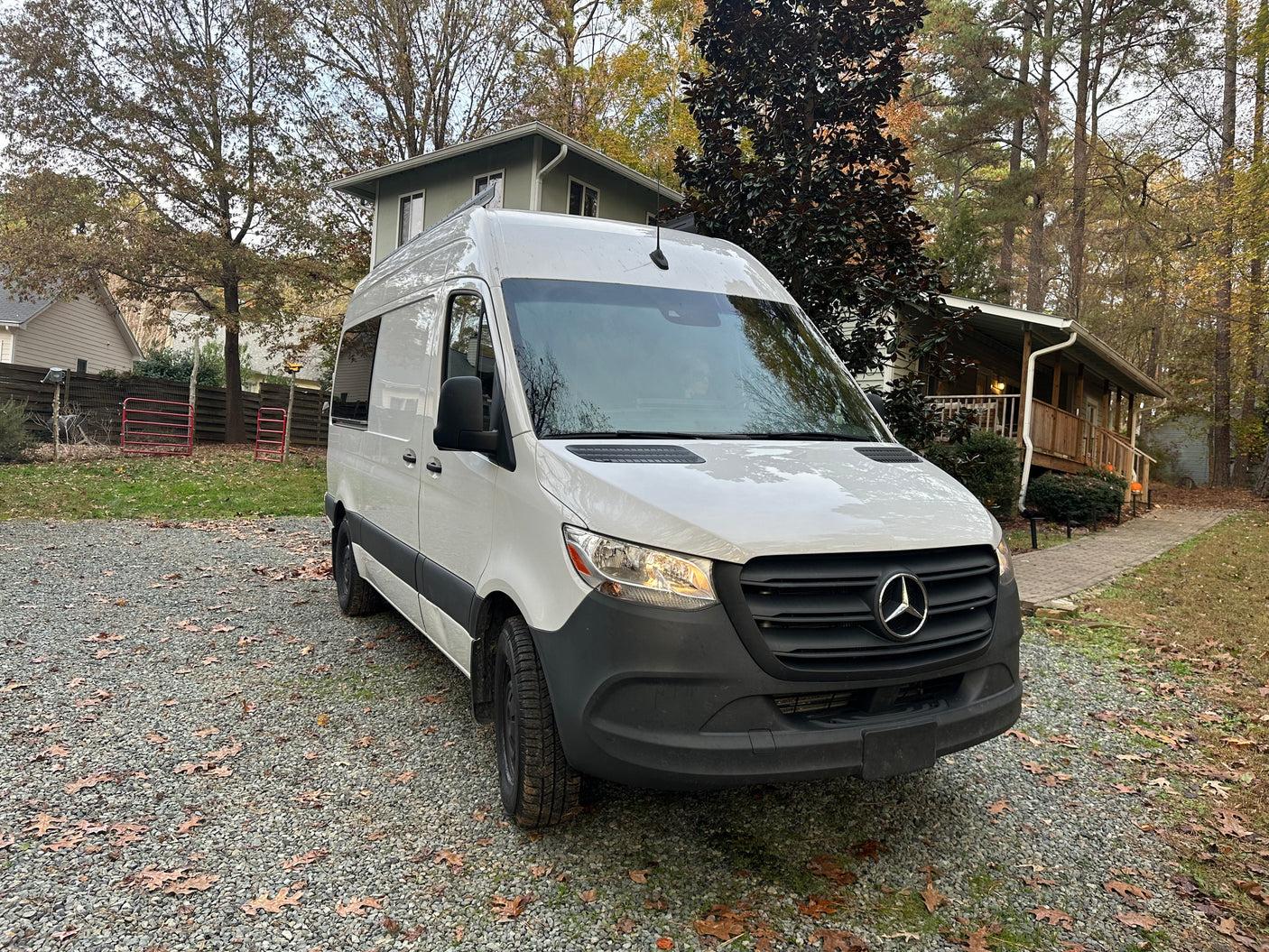 White Mercedes-Benz Sprinter van, ready for RV rental, parked on a gravel driveway with homes & trees.