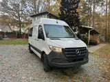 White Mercedes-Benz Sprinter van, ready for RV rental, parked on a gravel driveway with homes & trees.