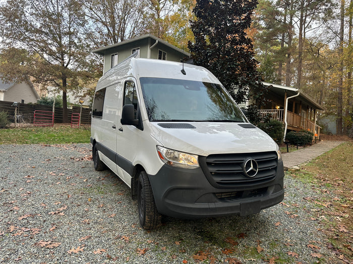 White Mercedes-Benz Sprinter van, ready for RV rental, parked on a gravel driveway with homes & trees.