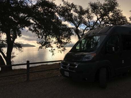 Experience serene travel with an RV rental: dark van by a tranquil lake, silhouetted trees at dawn.