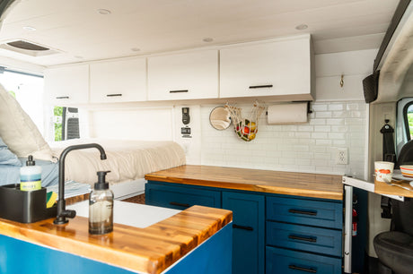 RV rental interior featuring a stylish kitchen with blue cabinets, wood counters, white subway tile, a sink, and a comfy bed.