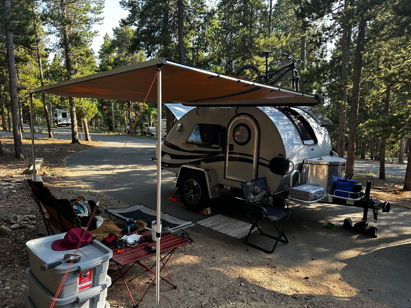 A modern teardrop camper with an extended awning at a peaceful forest campsite, showcasing an RV rental setup for outdoor adventure.