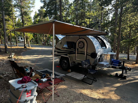 A modern teardrop camper with an extended awning at a peaceful forest campsite, showcasing an RV rental setup for outdoor adventure.