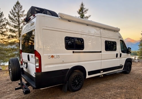  quilted bed & floral pillows. Arched windows reveal a beautiful lake & forest view.|Cozy bed in an RV rental looking out at a beautiful lakeside campsite with a sunset — photo 2