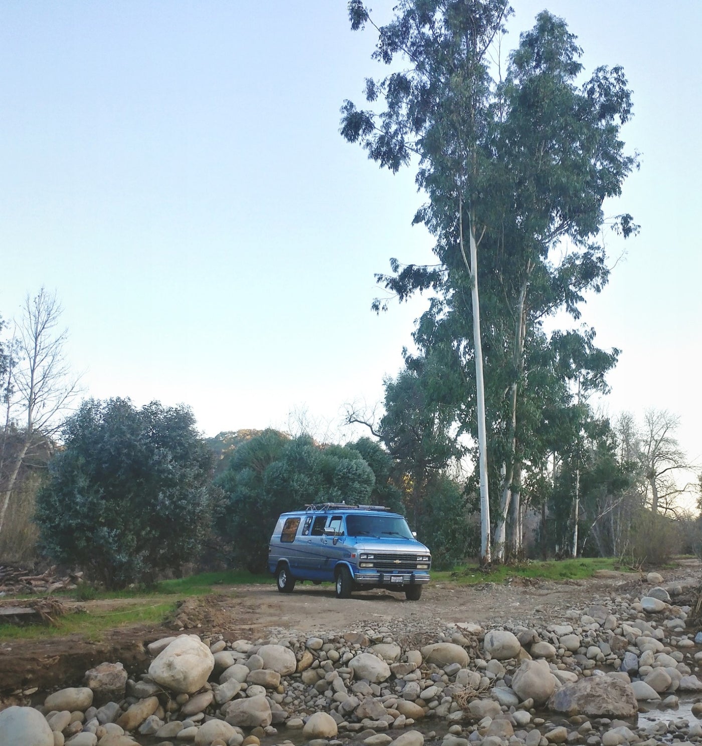 Blue RV rental parked by a rocky riverbed & tall trees in a serene natural setting. — photo 2