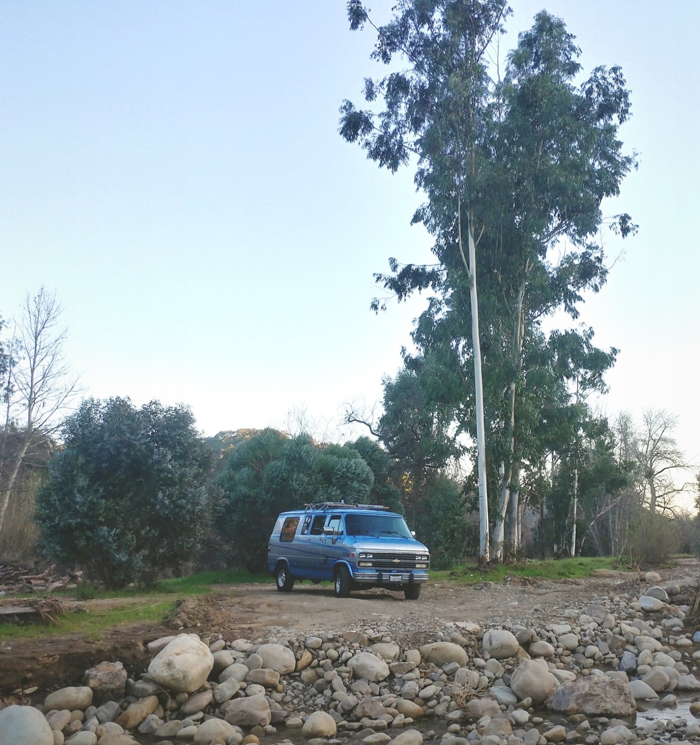Blue RV rental parked by a rocky riverbed & tall trees in a serene natural setting.