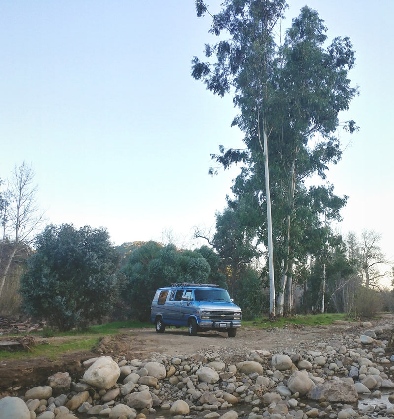 Blue RV rental parked by a rocky riverbed & tall trees in a serene natural setting. — photo 2