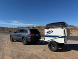 Grey SUV towing a white off-road RV rental trailer with a rooftop tent, ready for desert overlanding against mountains.