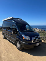 Dark blue Ford Transit camper van (RV rental) parked on a dirt path with a clear ocean view under a blue sky.