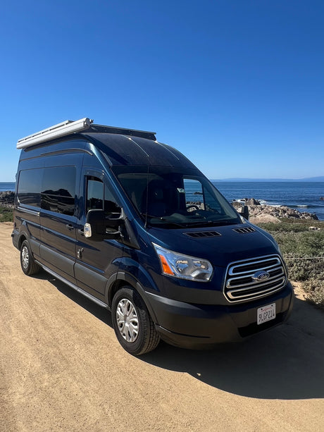 Dark blue Ford Transit camper van (RV rental) parked on a dirt path with a clear ocean view under a blue sky.