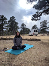 Woman practices yoga on a mat beside her RV rental, amidst pine trees under a cloudy sky.