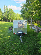 Enjoy nature with this compact RV rental! A white camper trailer is set up in a grassy field with chairs, surrounded by lush trees.