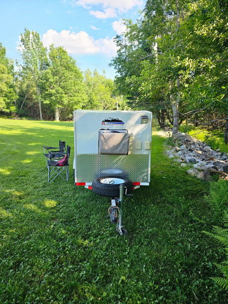 Enjoy nature with this compact RV rental! A white camper trailer is set up in a grassy field with chairs, surrounded by lush trees.