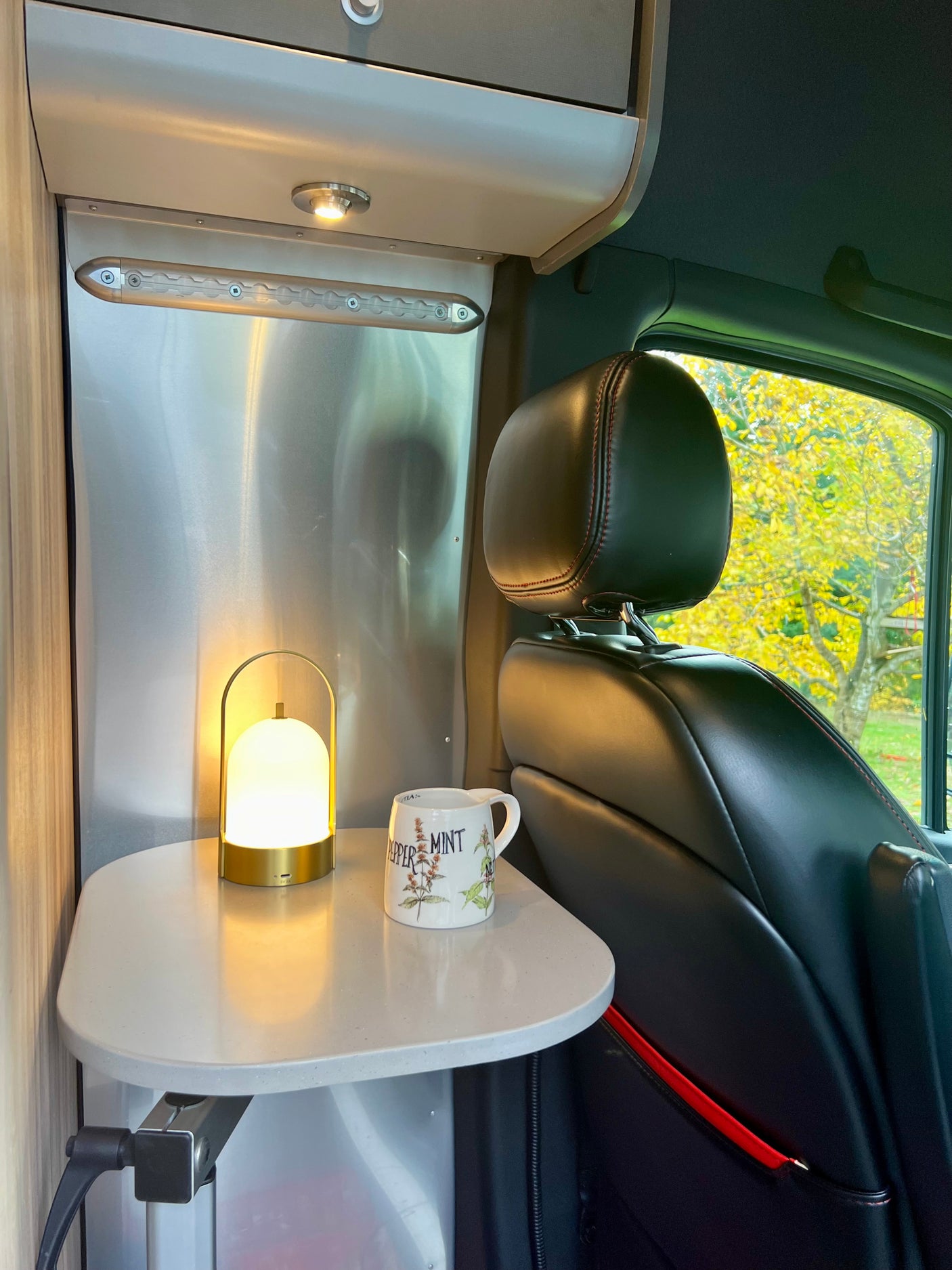 Cozy RV rental interior with a table, glowing lamp, peppermint mug, and vivid autumn tree view outside.