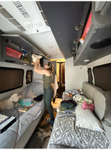 A woman organizes overhead storage in an rv rental, with two white dogs resting on beds and floor.