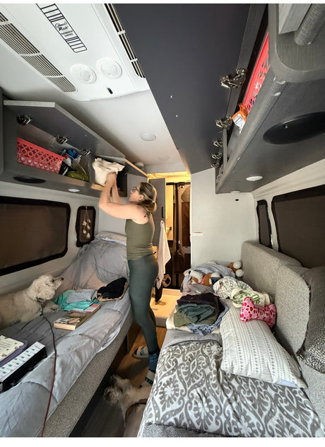 A woman organizes overhead storage in an rv rental, with two white dogs resting on beds and floor.