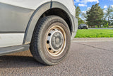 Close-up of a Ford van's tire and wheel (potential RV rental), parked on asphalt with grass and trees in the background.