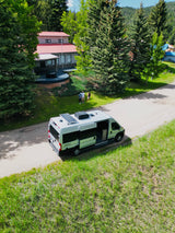 Your next adventure starts here! Spacious RV rental with solar panels on a scenic mountain road. People and house nearby.