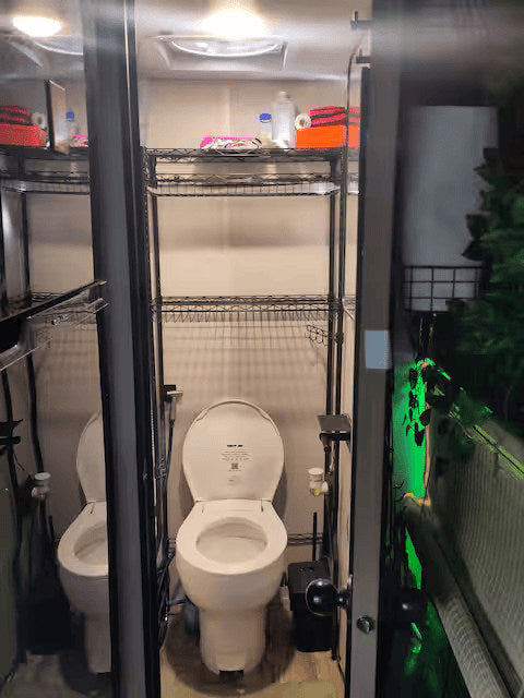 Inside an RV rental: a compact bathroom with a white toilet, open lid, and wire shelving above. Reflections suggest a narrow space.