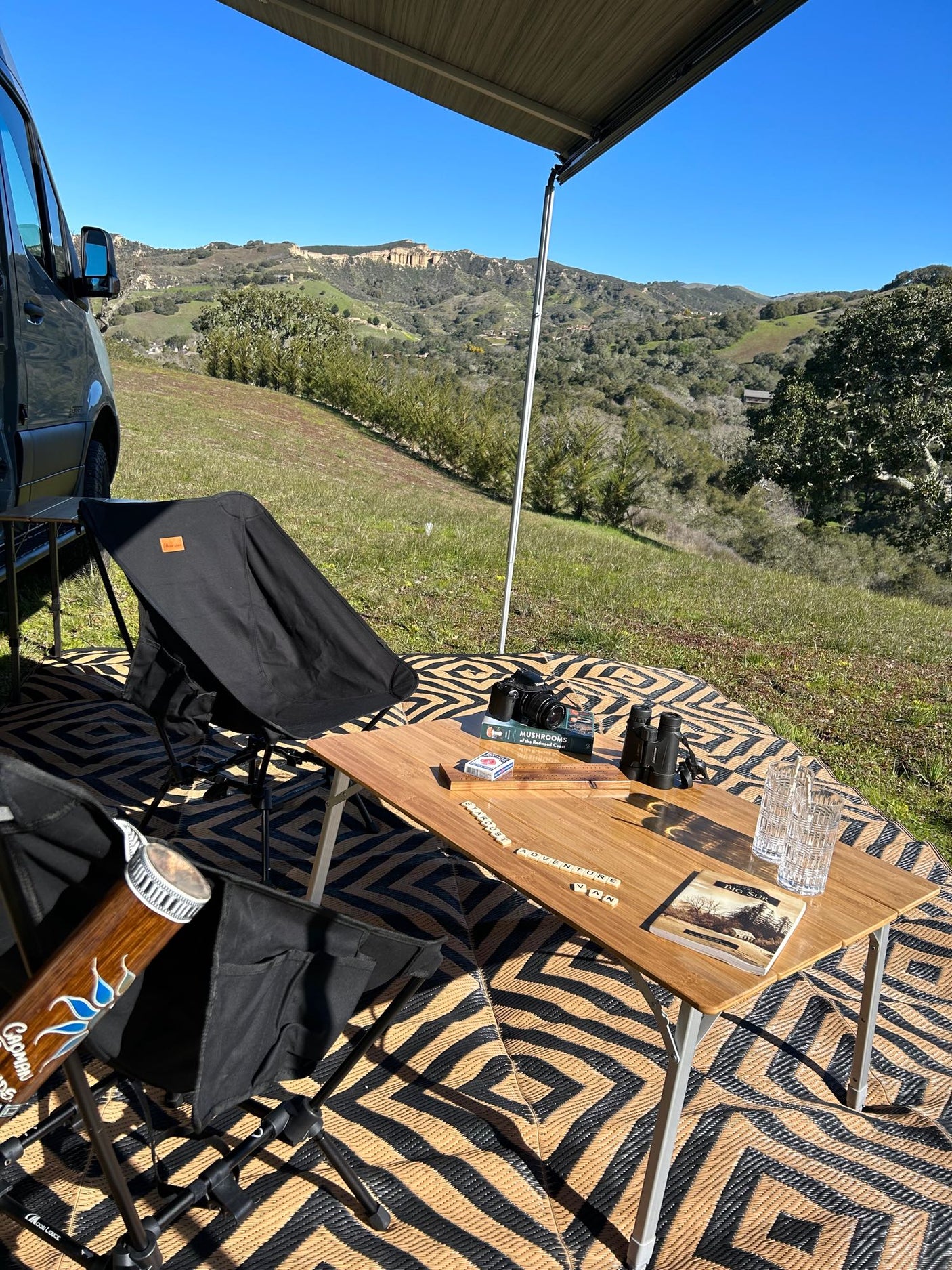 Enjoying an RV rental setup: camper van with awning, chairs, table with camera, binoculars, and games in a scenic valley.