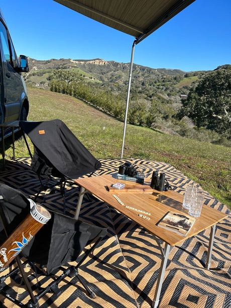 Enjoying an RV rental setup: camper van with awning, chairs, table with camera, binoculars, and games in a scenic valley.