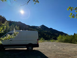 A white rv rental van is parked on a dirt lot, facing lush green mountains with a snowy peak under a bright blue sky.
