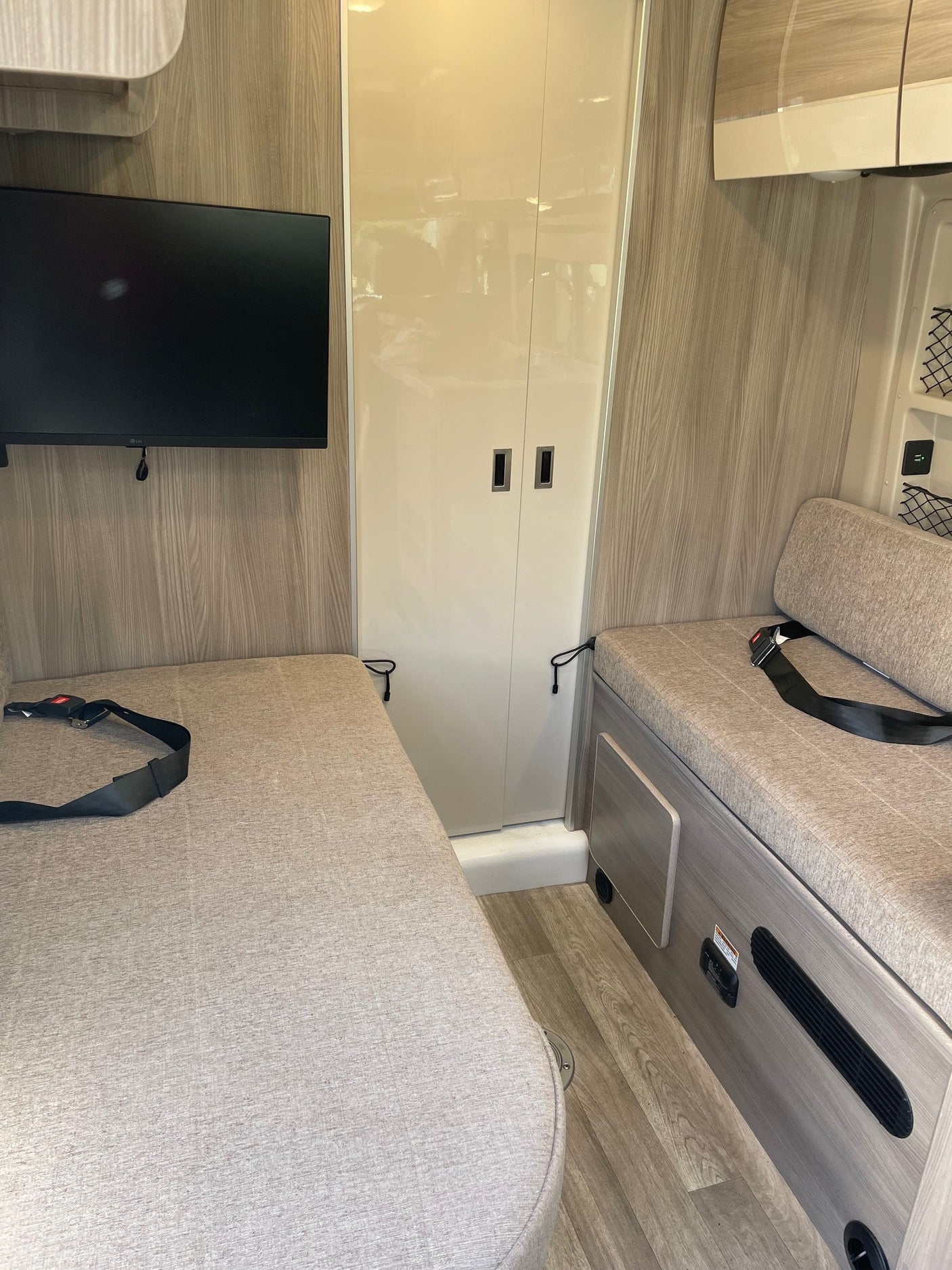 Interior of an RV rental: two cushioned benches with seatbelts, a wall-mounted TV, and a central cabinet.