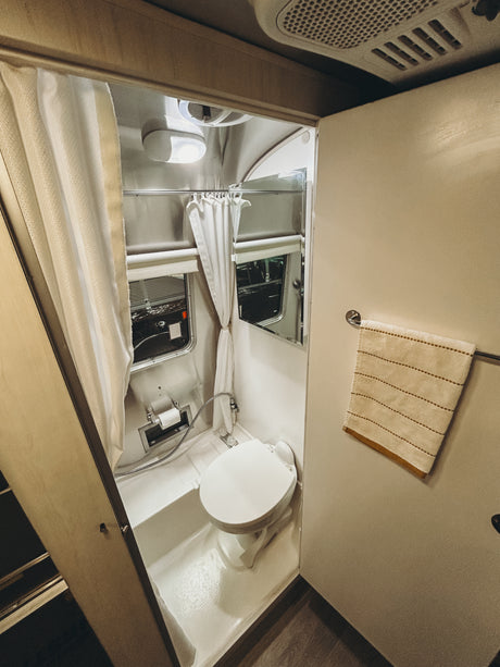 A functional RV rental bathroom showing a toilet, shower curtain, mirrored cabinet, windows, and a hanging towel.