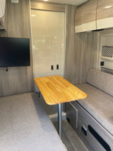 Modern RV rental interior with light wood-look paneling, a central table, facing gray benches, and a wall-mounted TV.