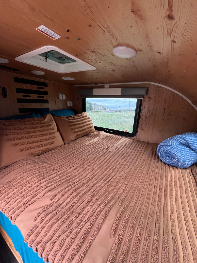 Cozy RV rental sleeping area with wood-paneled walls. Features a textured bed, pillows, and a window view of green fields. — photo 3
