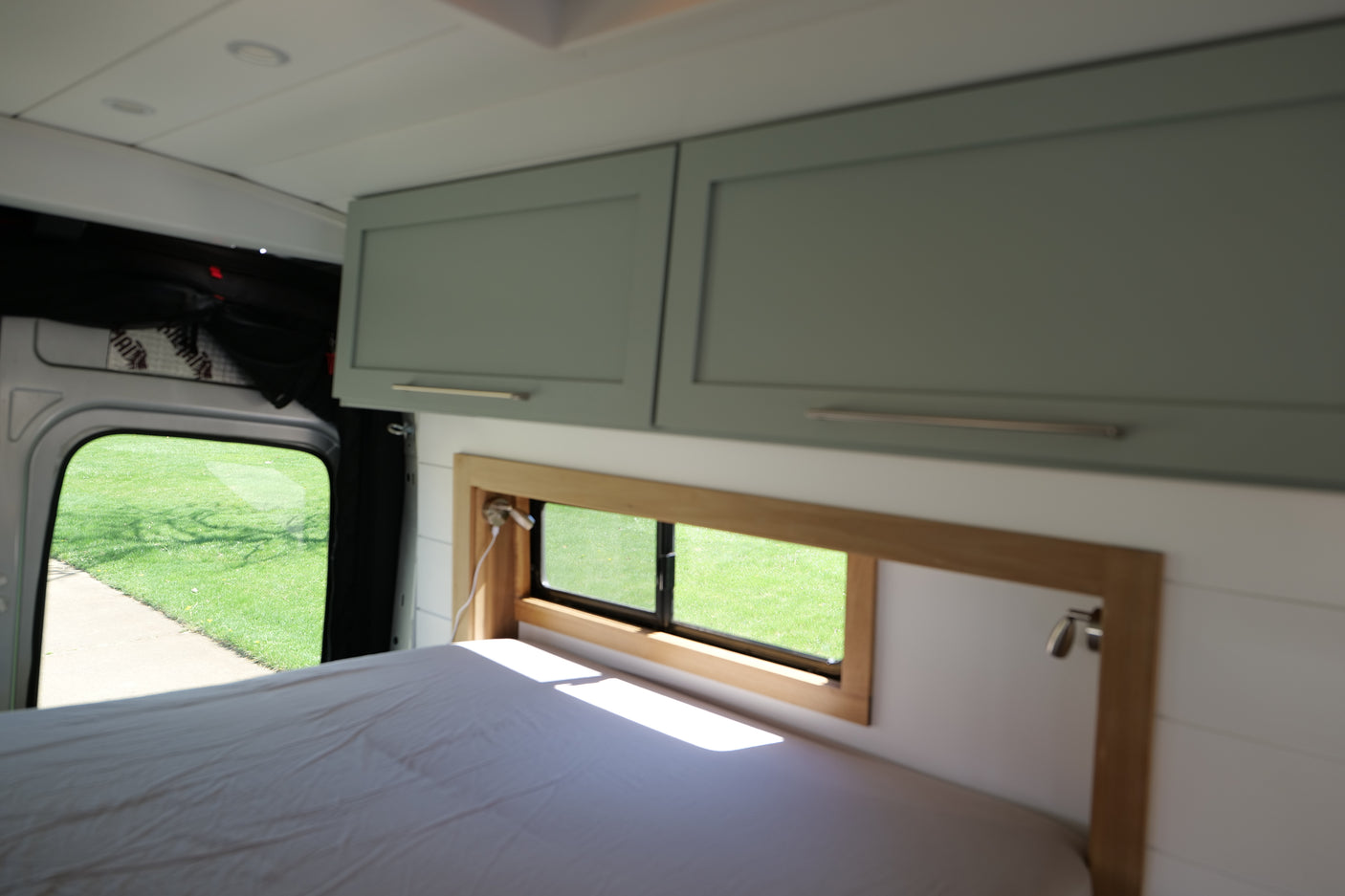 Experience comfort in this RV rental: a cozy bed, mint green cabinets, and large windows offering views of green grass outside.