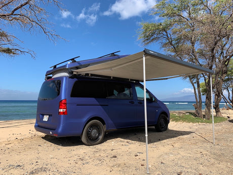 Blue pop-top camper van with extended awning at a sunny beach. Great RV rental for oceanfront adventures!