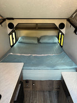 Cozy RV rental bedroom: large bed with light blue bedding, side windows, overhead shelves, and integrated speakers.
