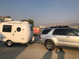 An RV rental, a white teardrop camper, hitched to a silver SUV, parked at a general store with a commercial sign and mountains.
