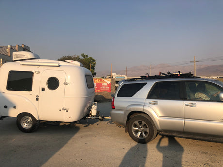 An RV rental, a white teardrop camper, hitched to a silver SUV, parked at a general store with a commercial sign and mountains.